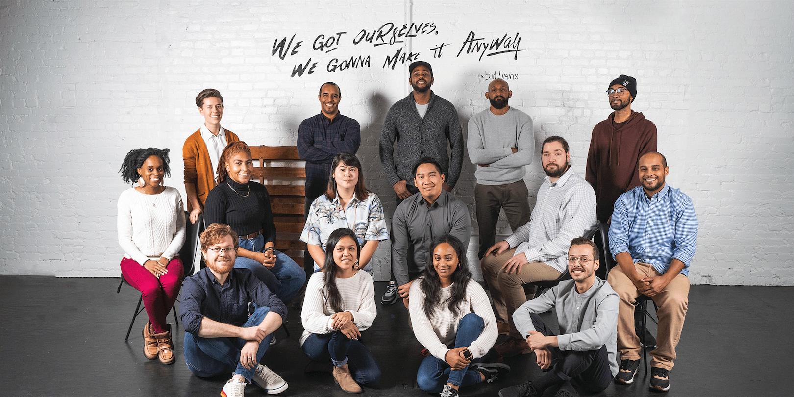 Image of the Ghost Note staff. Some staff are standing, some are seated in chairs and some are sitting on the floor. There is also a white wall behind them and a wooden bench.